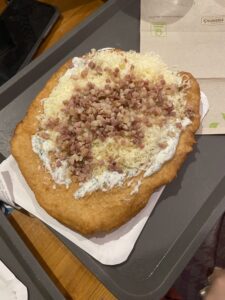 Lángosz to węgierska ikona street foodu - smażone ciasto drożdżowe, chrupiące z zewnątrz i miękkie w środku. W swojej prostocie jest doskonały! Tradycyjnie podaje się go z kwaśną śmietaną, tartym serem, a czasem też ze skwarkami lub cebulą. Dziś wersji jest znacznie więcej - od klasycznych po nowoczesne, z czosnkiem, szynką, a nawet na słodko.